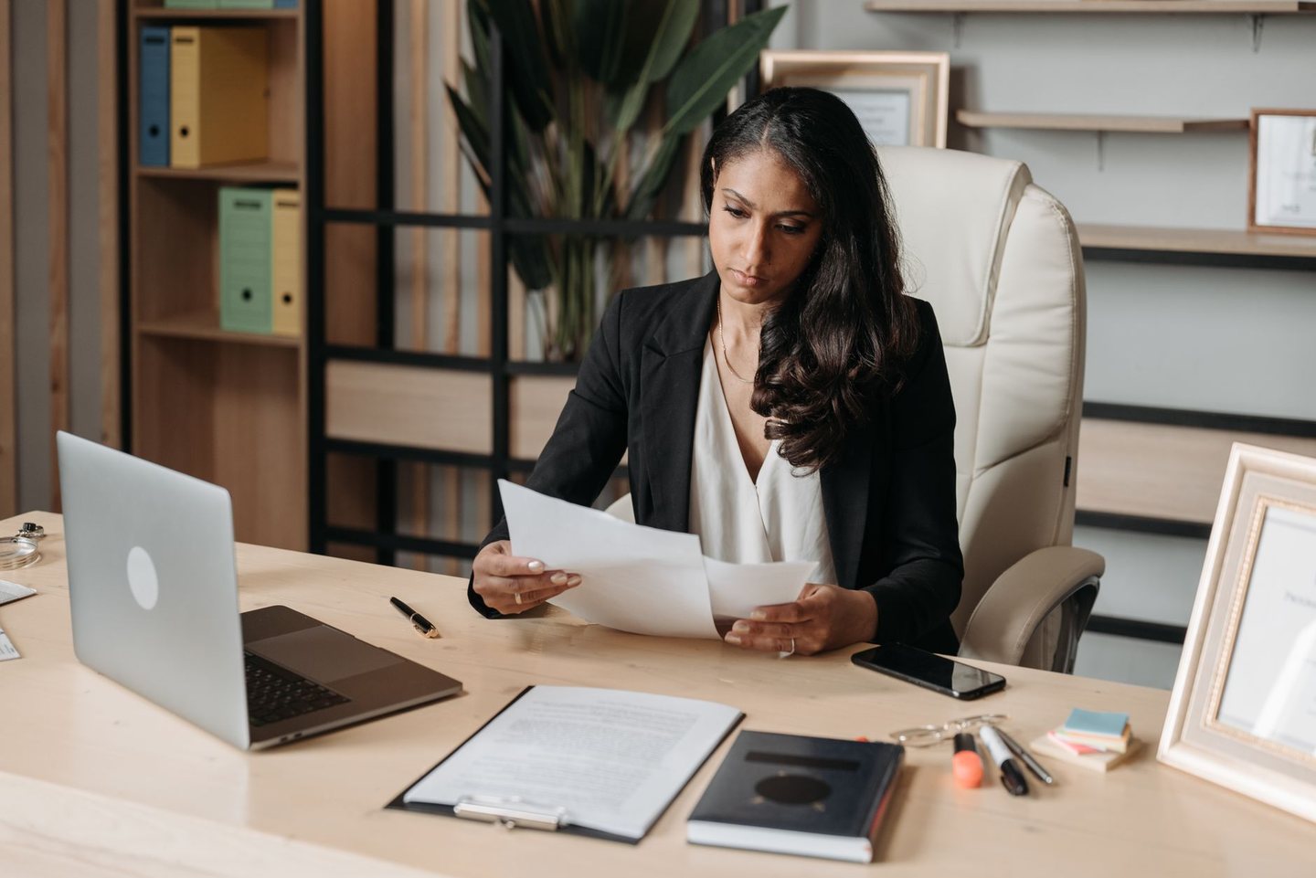 A contracts manager at her desk reading two pages, a laptop and a clipboard beside her